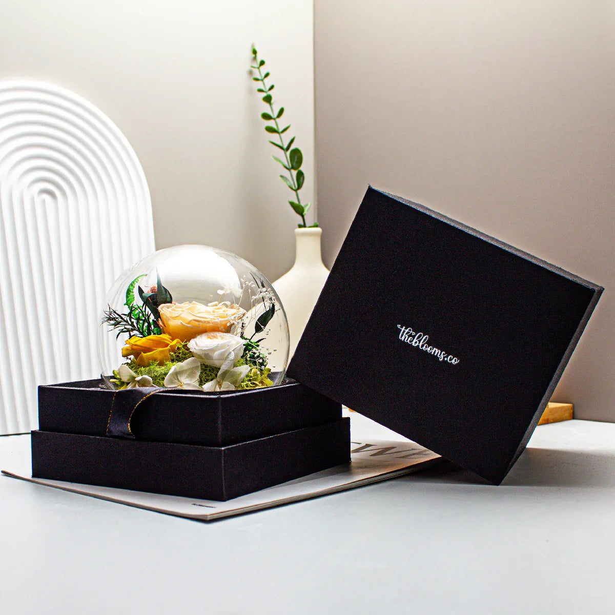 The Secret Garden preserved floral arrangement in a glass dome featuring yellow and white roses with green foliage, displayed in a black theblooms.co gift box.